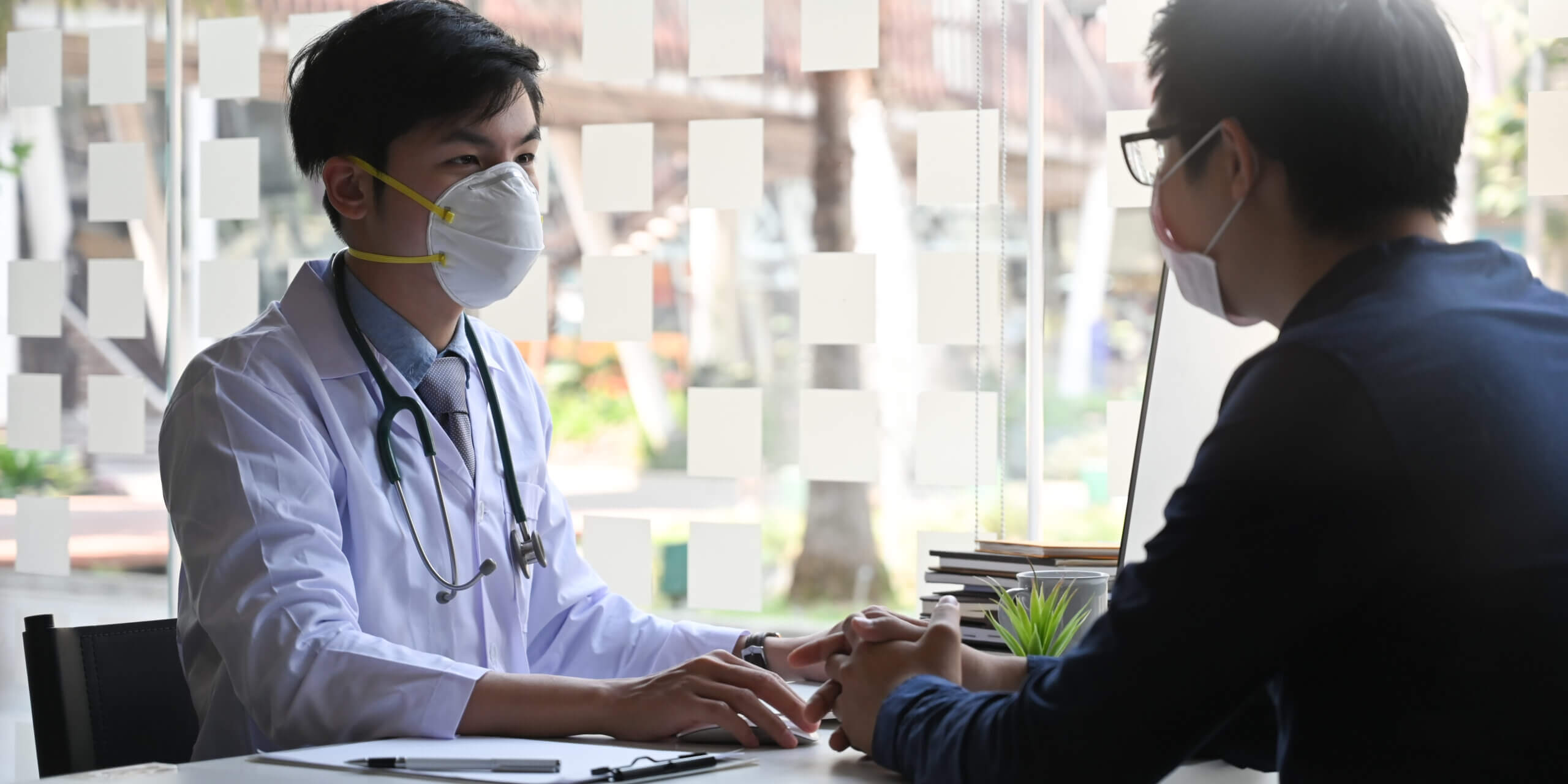 Doctor talking/explaining with his patient while sitting together as working desk over modern examination room as background.
